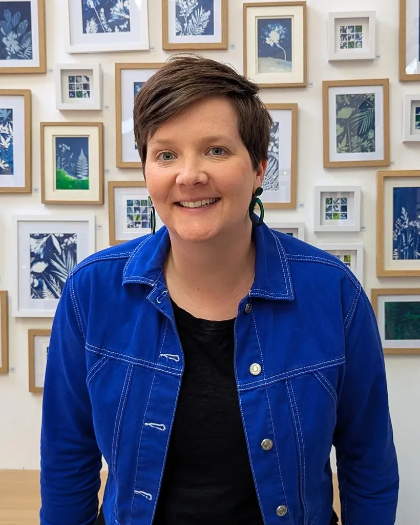 Artist Fiona Clabon wearing a blue jacket, smiling in front of a gallery wall filled with framed cyanotype artworks